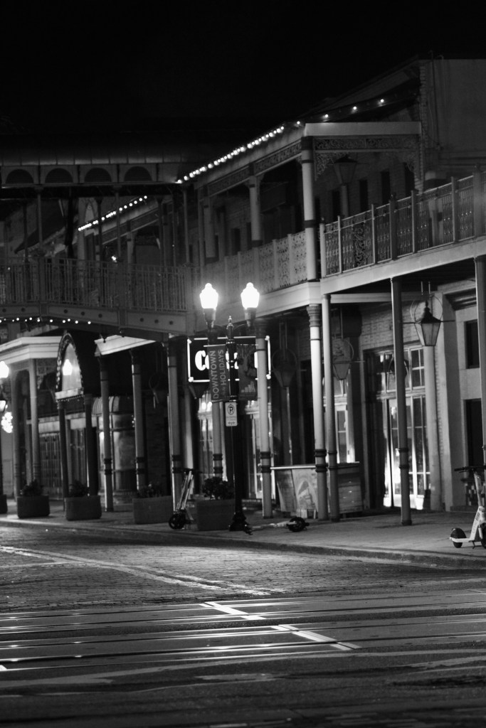 A black and white night scene of a historic street featuring decorative buildings, street lamps, and a small sign partially visible, creating a quiet atmosphere.